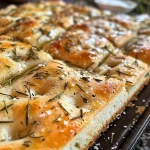 Juicy focaccia bread with a crispy crust, garnished with rosemary and sprinkled sea salt, captured in bright lighting.