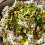 A detailed image of whipped herb feta adorned with lemon zest and fresh herbs, with a few whole pistachios on top.