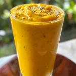 Close-up of a vibrant mango peach smoothie in a clear glass, garnished with a slice of fruit.