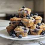 ['Close-up side view of a blueberry Greek yogurt muffin with a golden-brown top.', 'Juicy blueberry Greek yogurt muffins stacked on a plate, showing the moist texture.', 'A single blueberry muffin with fresh blueberries scattered around it, highlighting its fluffy interior.', 'Side view of a blueberry muffin, showcasing its crumbly texture and plump blueberries.']