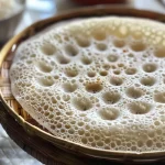 ['Close-up of a fluffy Appam pancake with a soft texture, garnished with coconut.', 'Side view of golden-brown Appam pancakes stacked on a plate, showcasing their fluffy interior.', 'Juicy, steaming Appam pancakes presented artistically with a drizzle of coconut milk.', 'A delicious Appam pancake with a soft, spongy surface, highlighted by its rich color.']